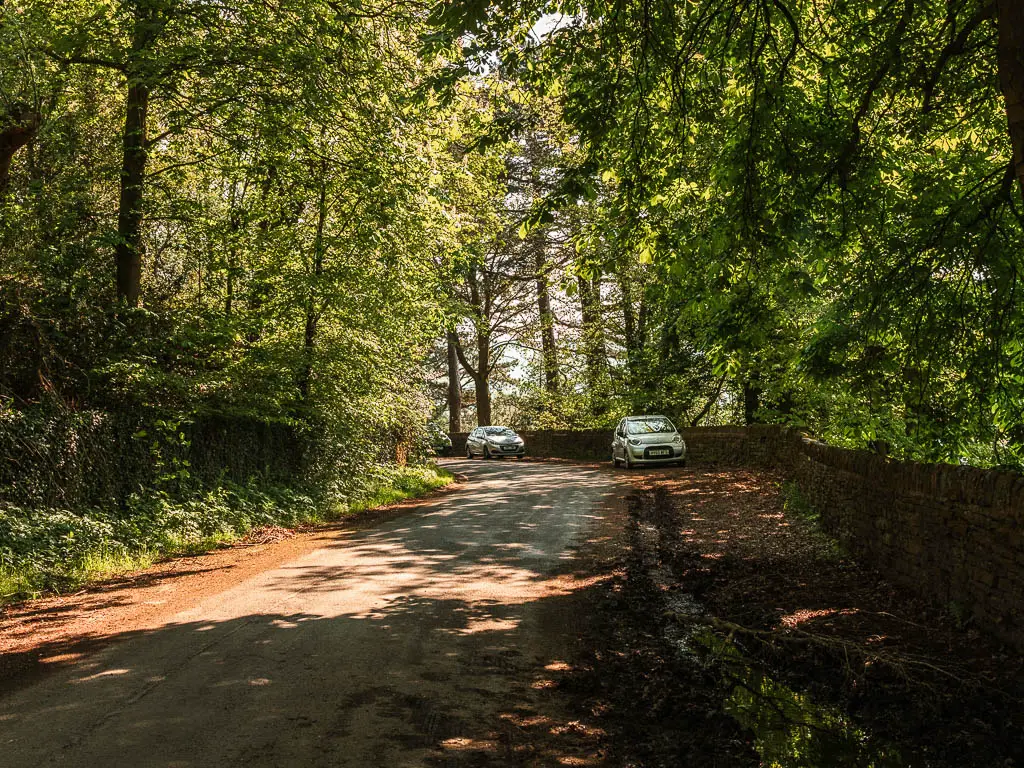 A road with two parked cars, next to the Agden Reservoir. the road is lined with green leafy trees. Rays of light are shining down through the trees.