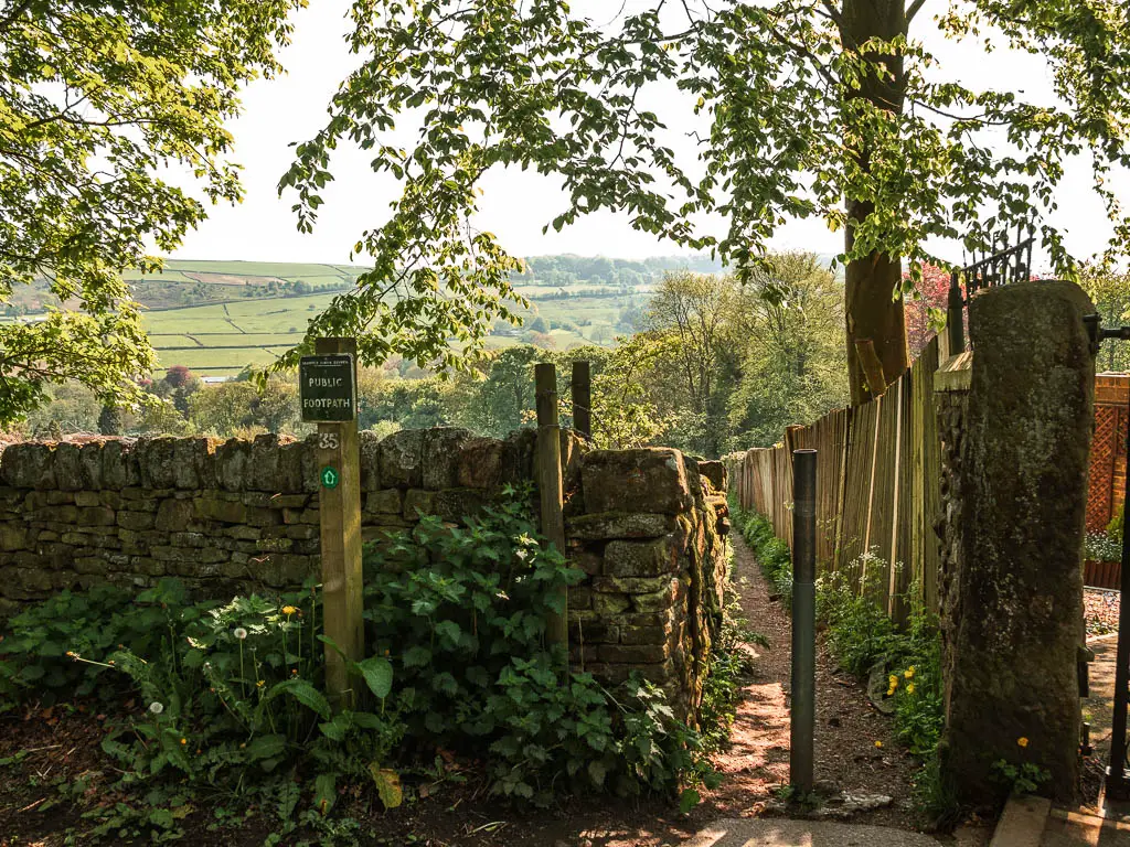 A gap leading to a thin trail, with a stone wall on the left and wooden fence on the right. there is a footpath sign at the entrance.