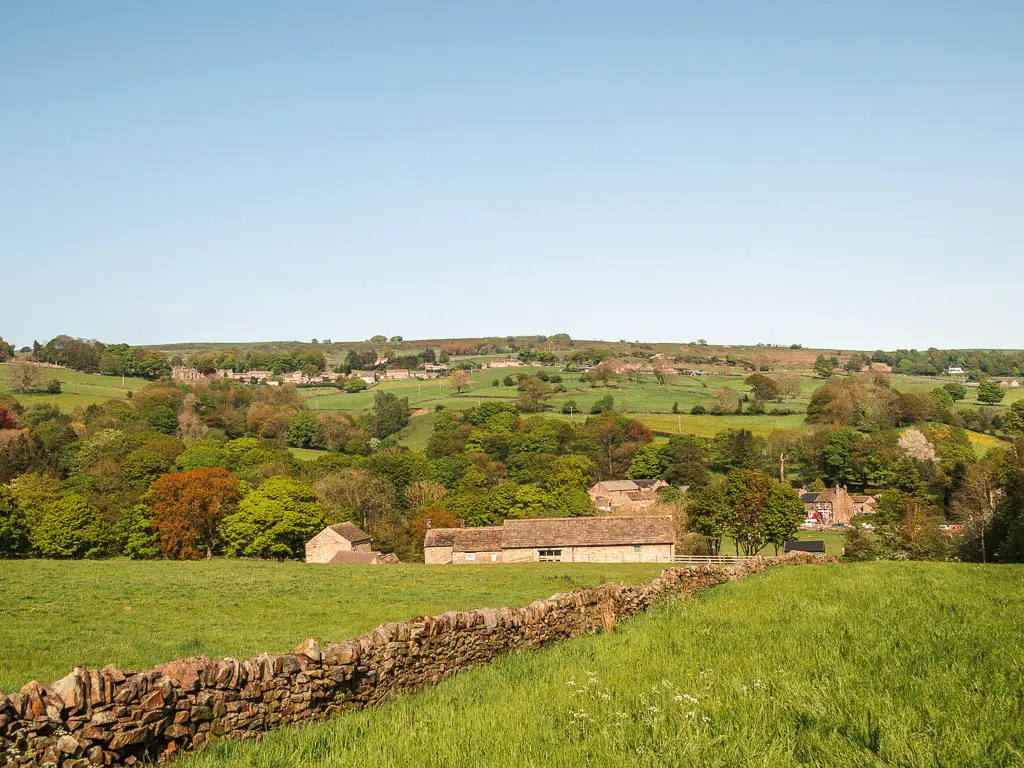 Looking across the fields of green grass, with a stone wall running down the middle of it, and a view beyond of the rooftops of houses, and woodland trees up the hill.
