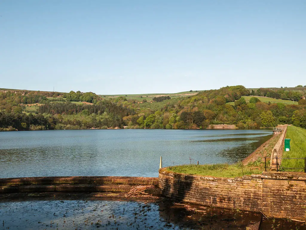 Looking across the Agden Reservoir at the start of the walk around it. There is a hill covered in green leafy trees on the other side.