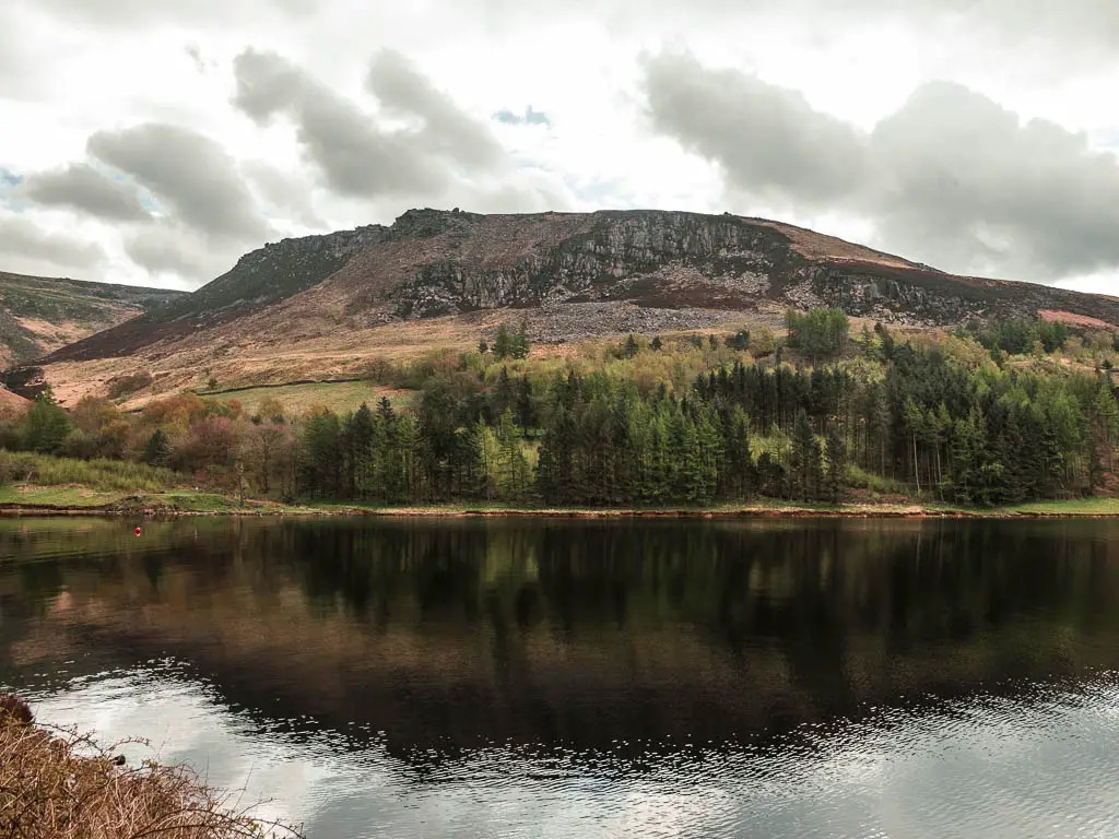A big hill on the other side of the reservoir, casting a shadow down on it. There is a mass of trees at the bottom of the hill.