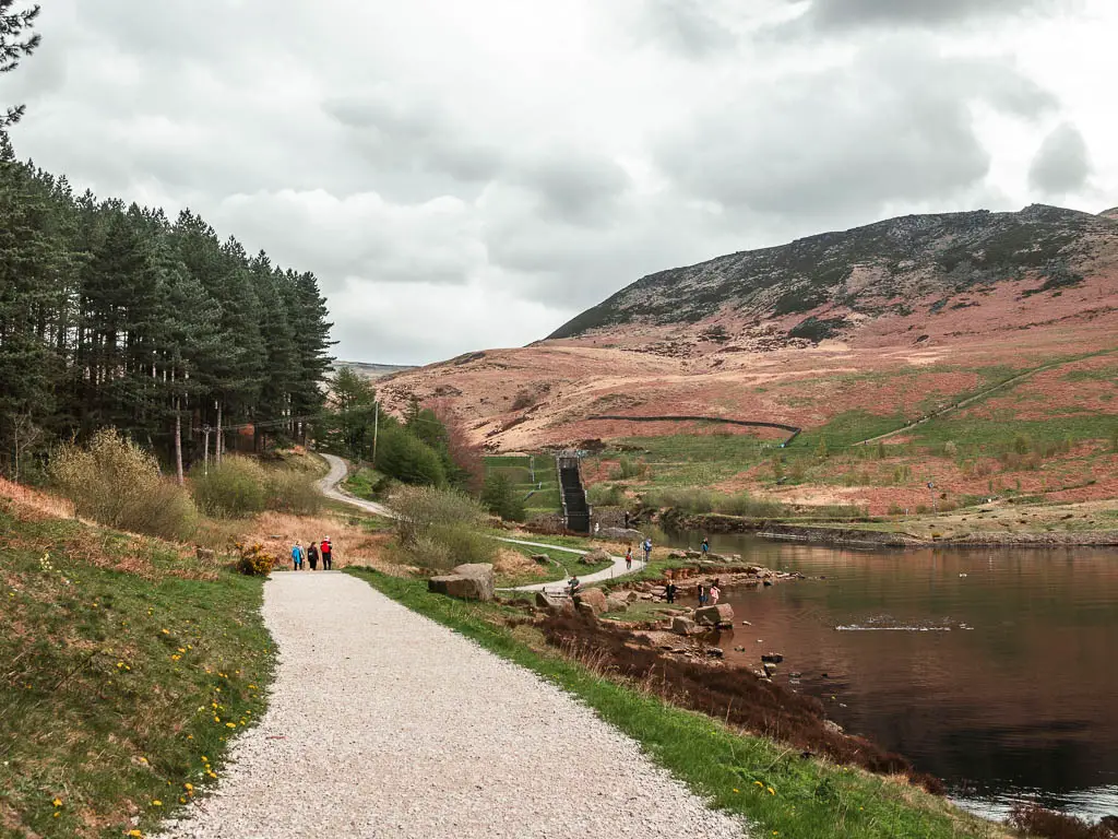 A gravelly path leading straight, with the reservoir to the right, near the end of the Dovestone section of the walk. There are a few people walking on the path ahead, and a big hill rising up on the other side of the reservoir to the right.