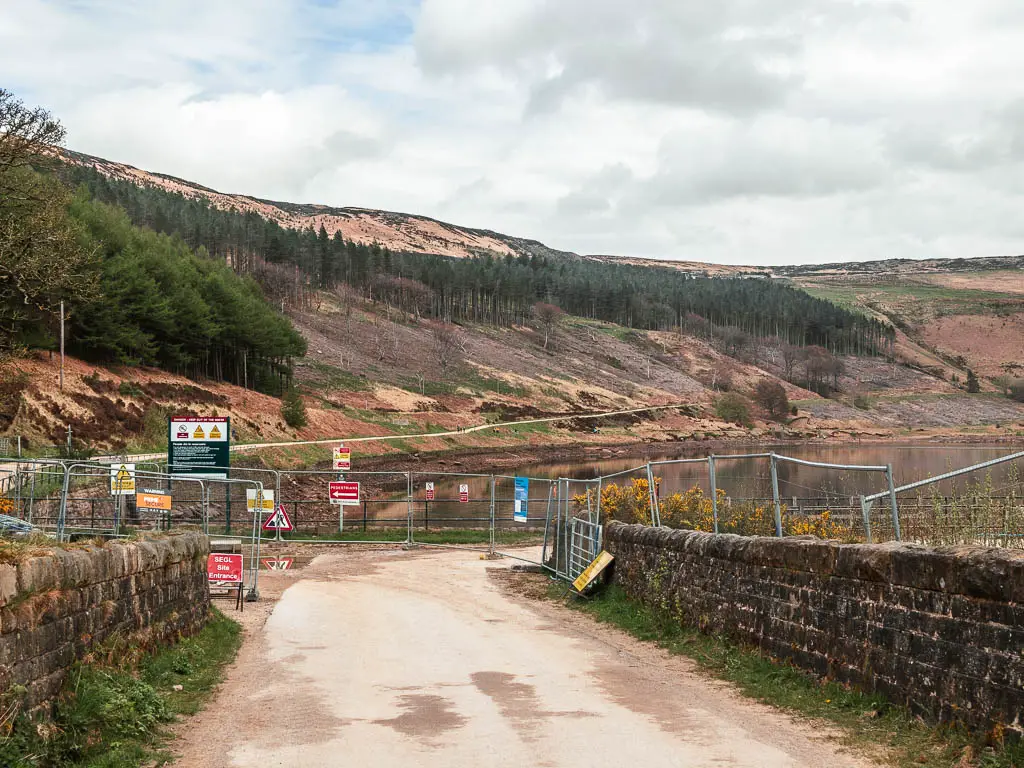 A wide path lined with stone walls and metal barrier fences ahead, with Yeoman Hat reservoir on the other side.