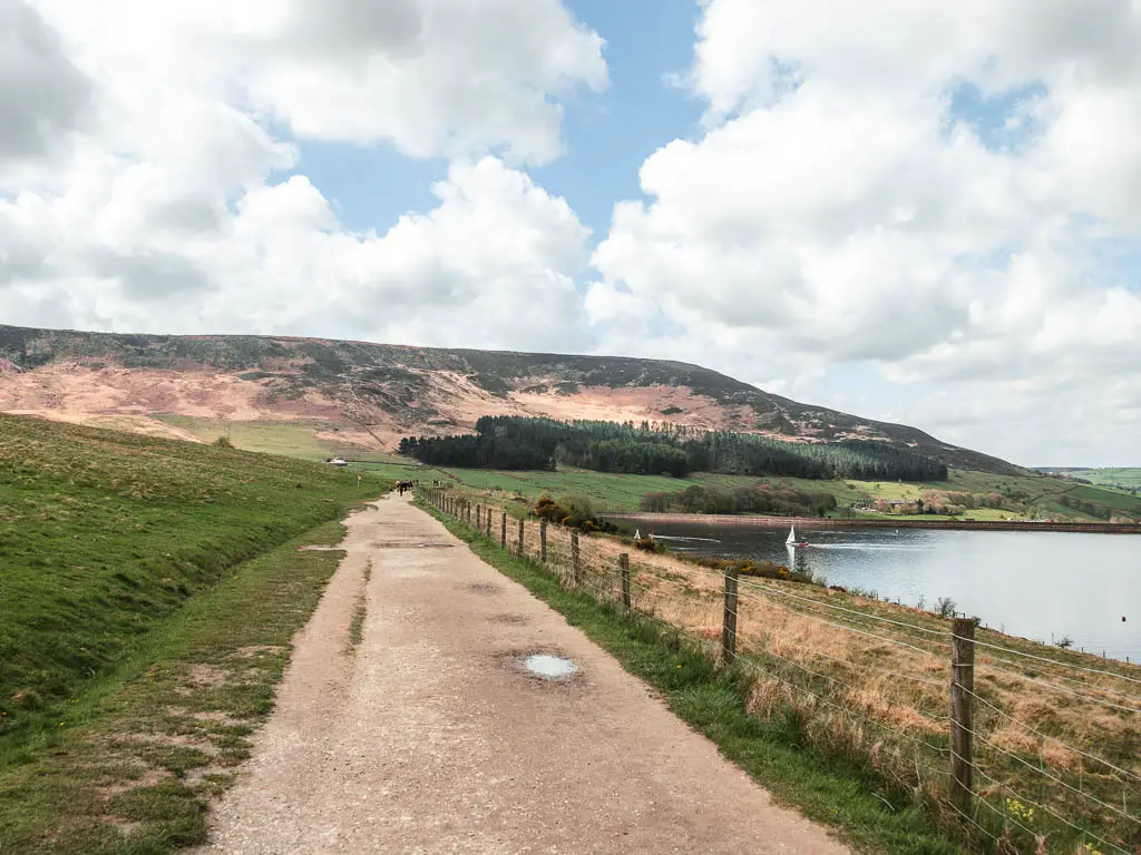 A wide path with a grass hill to the left and a wire fence pin there right, with the water to the right of the fence. The path leads straight ahead with a hill ahead in the distance.