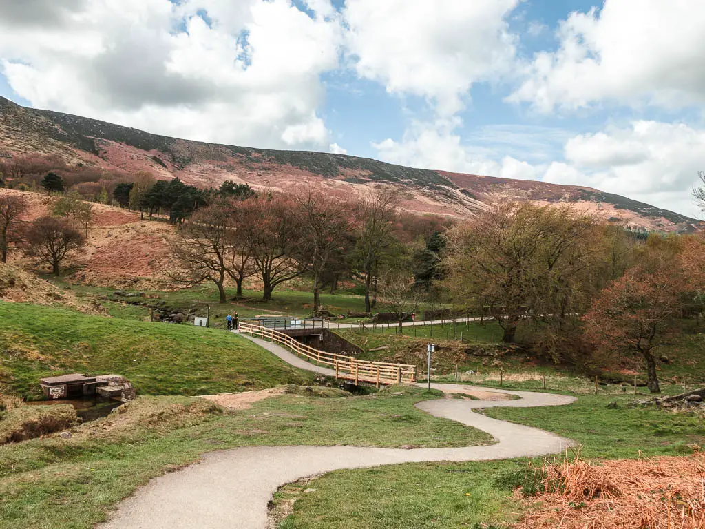 A path snaking ahead through the grass, with some trees ahead and a hill beyond them.