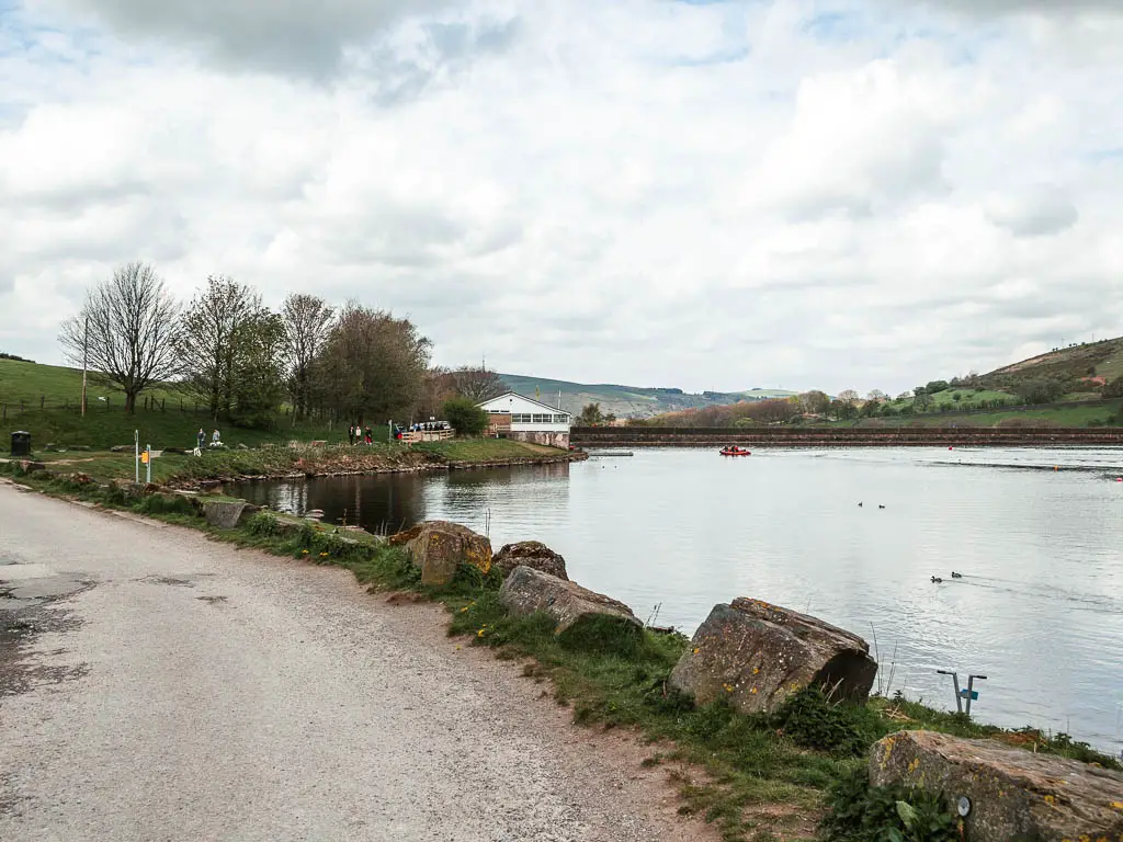 A gravel road curving left, lined with rocks on the right, and the reservoir on the right side. There is a boat house ahead on the edge of the water.