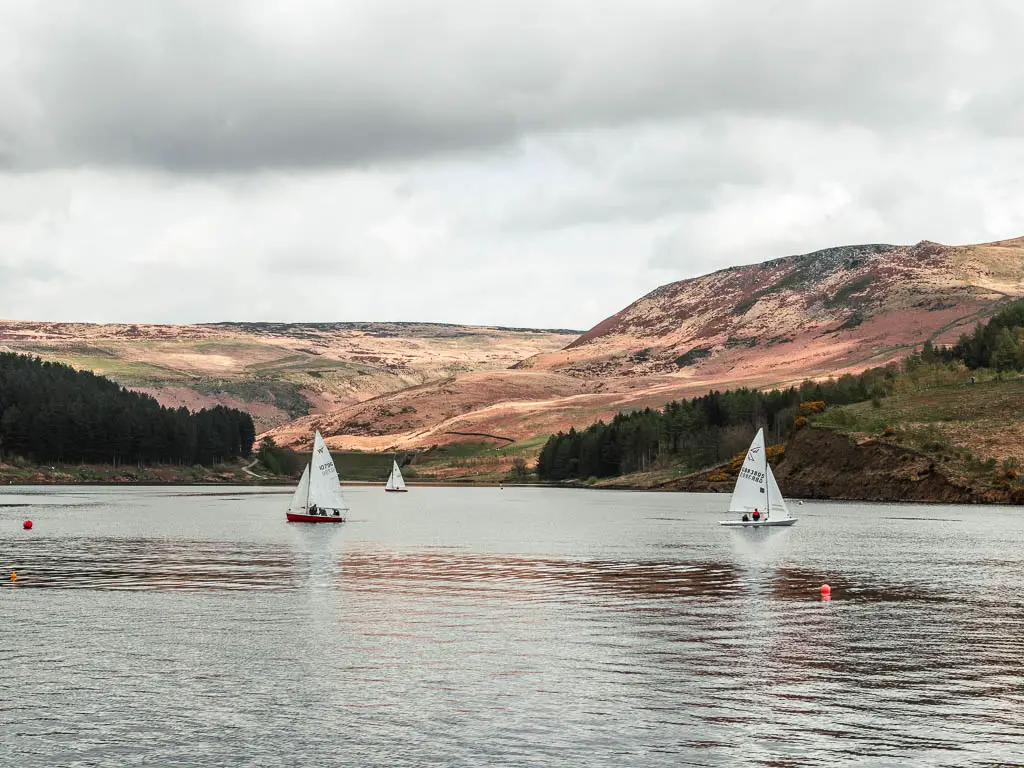 A few sailboats on the water of dovestone reservoir in the Peak District, on the circular walk around it. there are undulating hills on the other side.