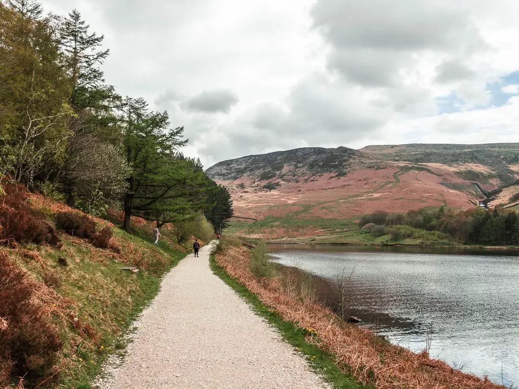 A well maintained path along the left side of the dovestone reservoir, with a person walking along it, in the distance. There are trees up the green grass bank on the left side of the path, and moorland hills on the other side of the reservoir.