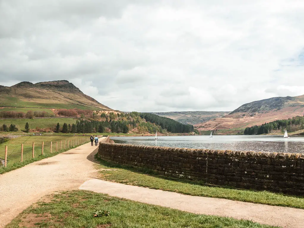 A well maintained path running around the corner of the dovestone reservoir, with two people walking on it straight ahead. There is a stone wall around the reservoir, and moorland hills undulating ahead in the distance.