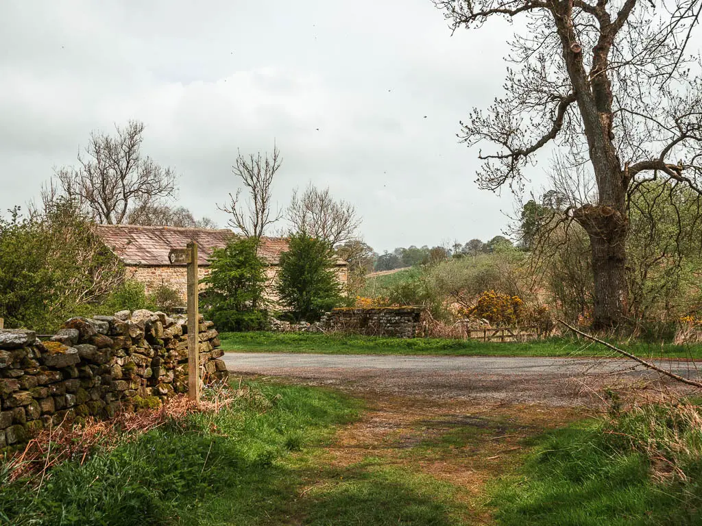 The grass path leading onto the side of a road, with a stone wall and wooden trail signpost on the left. There is a stone hut building on the other side of the road to the left.