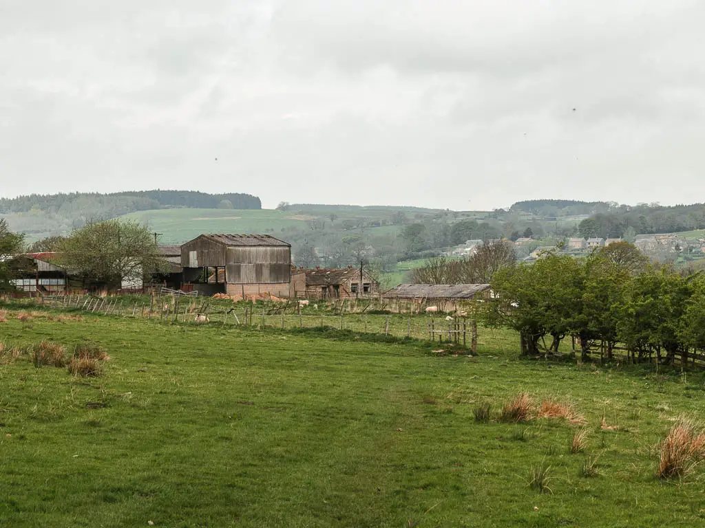 A grass field with a fence ahead, and a field on the other side. There are farm buildings on the other side of the second field.