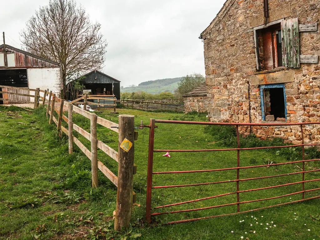 A wooden fence and metal gate corner with a stone budding on the right and farm sheds ahead.