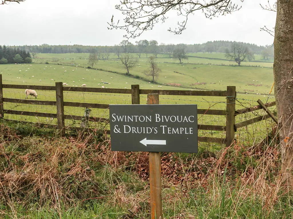 A grey sign with white writing saying 'Swinton Bivouac and Druid's Temple' and an arrow pointing left to walk to it. There is a wooden fence and field behind the sign.