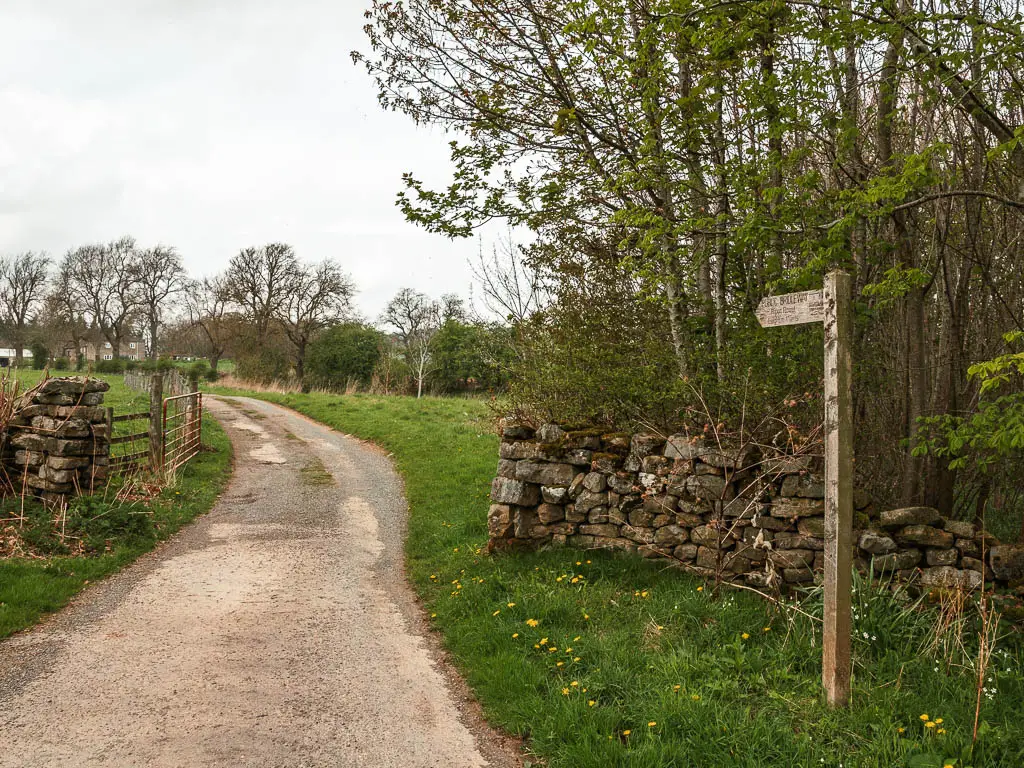 A road leading through a gap in the stone wall, and a wooden trail signpost on the right, on the walk towards Druid's Temple.