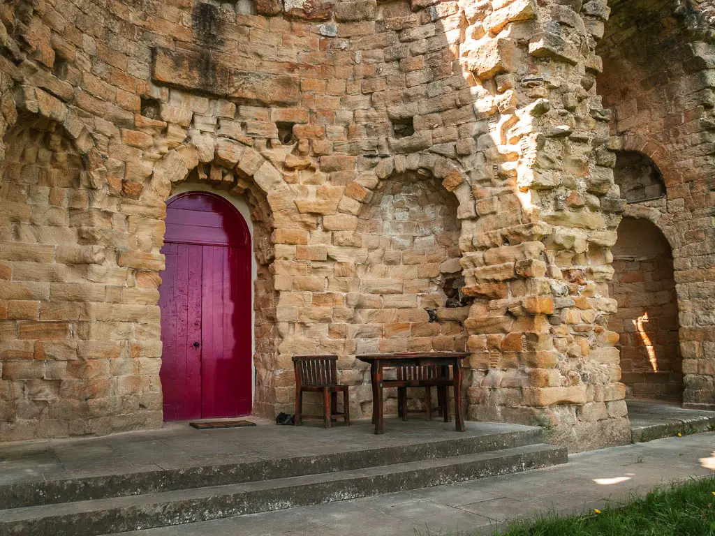 A red door in the stone archway wall, with a dark brown wooden table and chairs next to it.