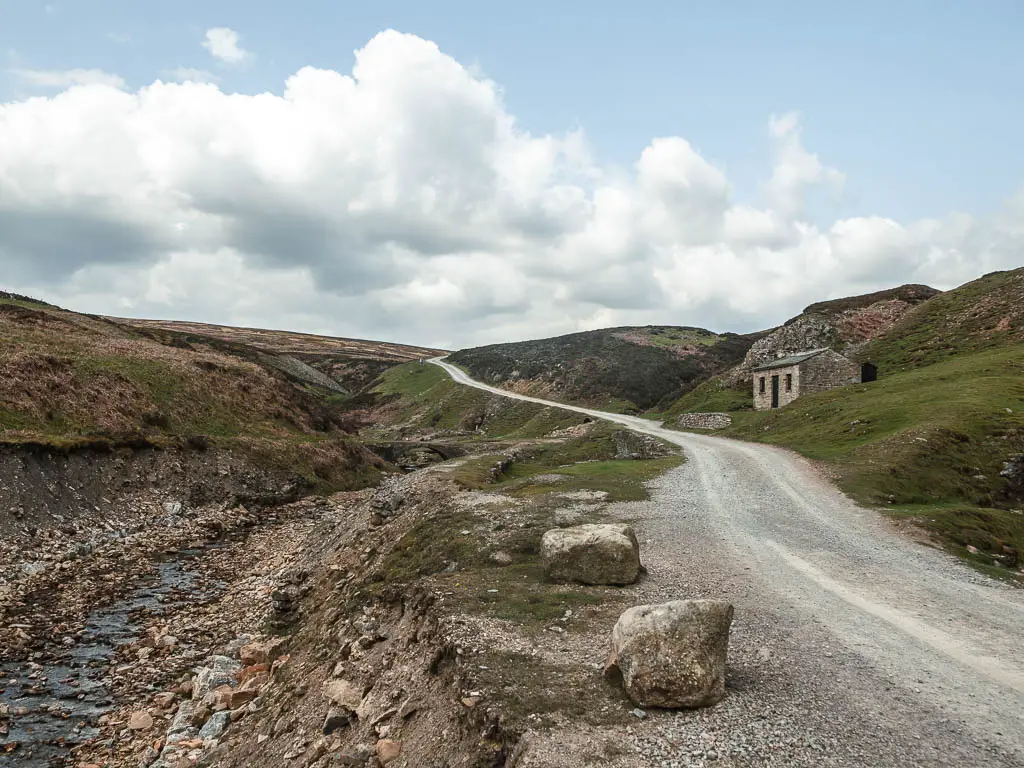 A grey gravel road, winding along the small valley hill ahead, with the river to the left, and an old mine building of Old Gang Smelt to the right, part way through the walk.