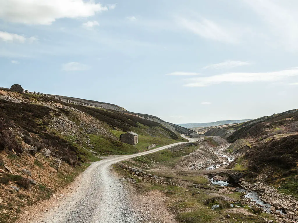 A grey gravel path winding ahead along the bottom of the hill, on the walk towards Old Gang Smelt. There are old mine buildings ahead next to the trail, and a river below to the right.