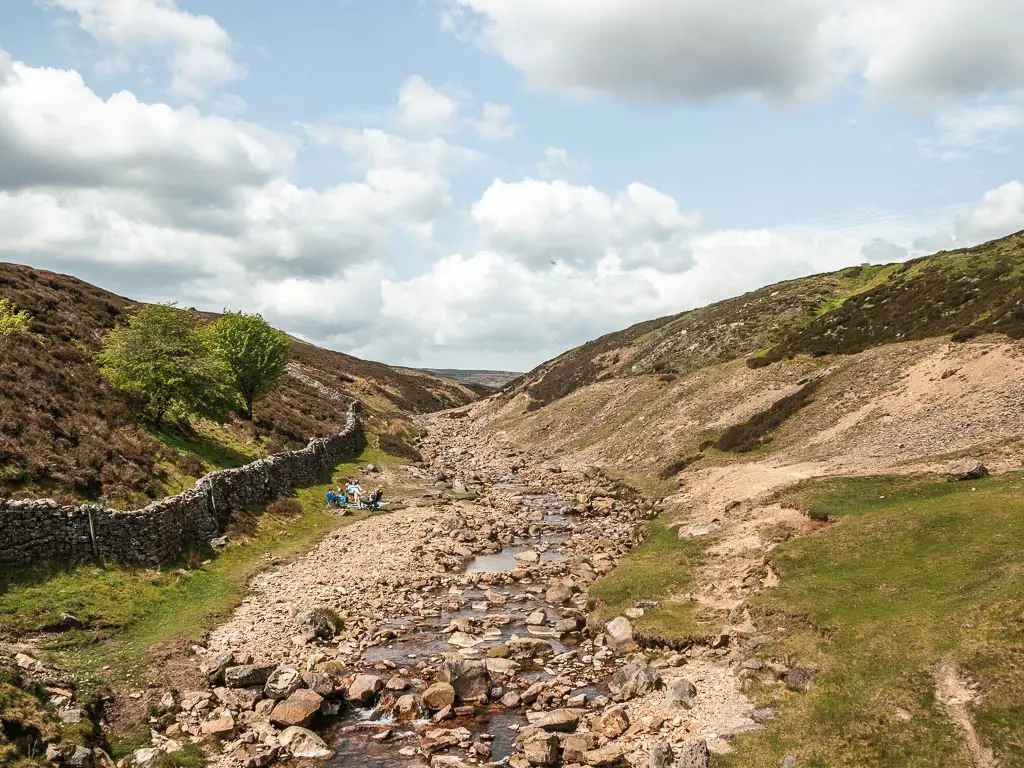 A rock filled river flowing through the valley ahead, at the start of the walk towards the old gang smelt mill. There are people sitting on picnic benches ext to the river.