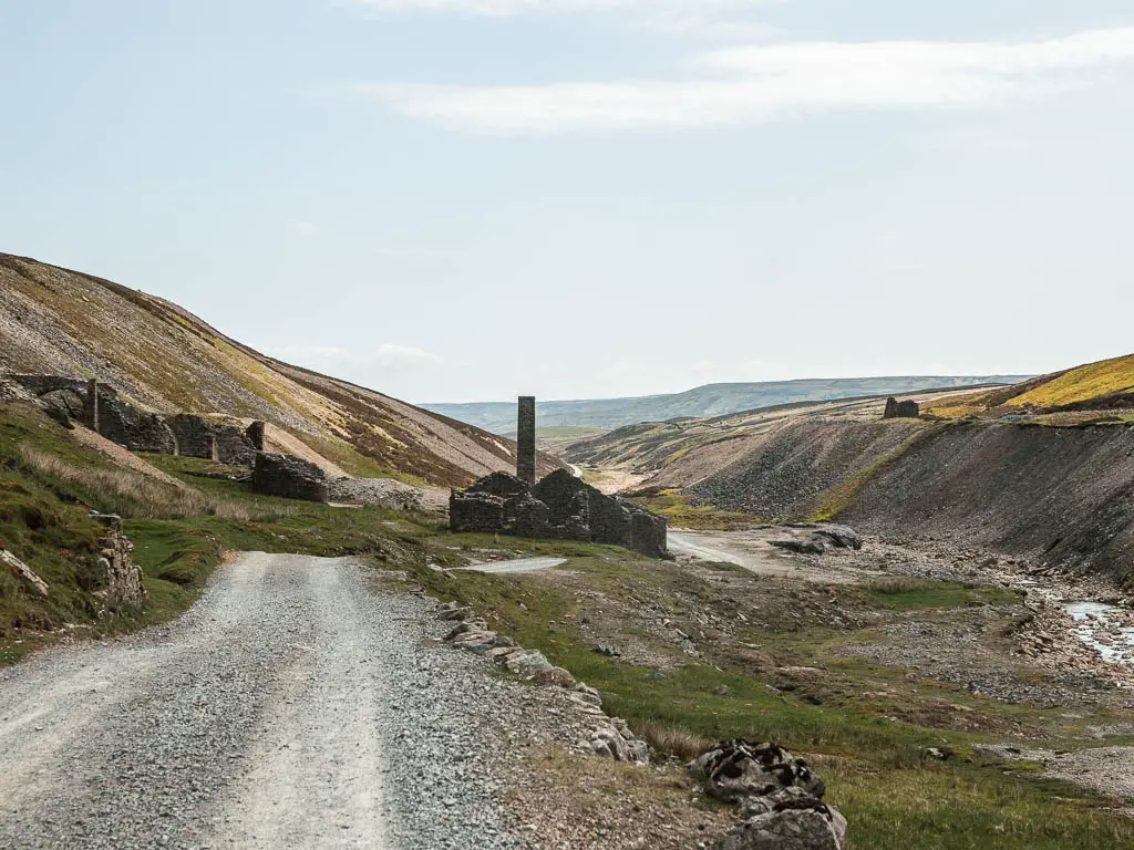 A grey gravel road leading straight towards the ruins of the old gang smelting mill buildings at the bottom of the valley, part way along the circular walk.