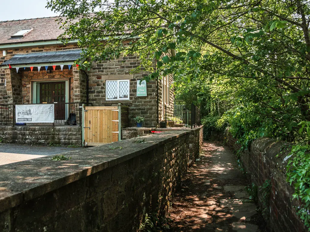 A small path lined with short walls. There are green leafy trees overhanging the right wall, and a small cafe on the other side of the left wall.