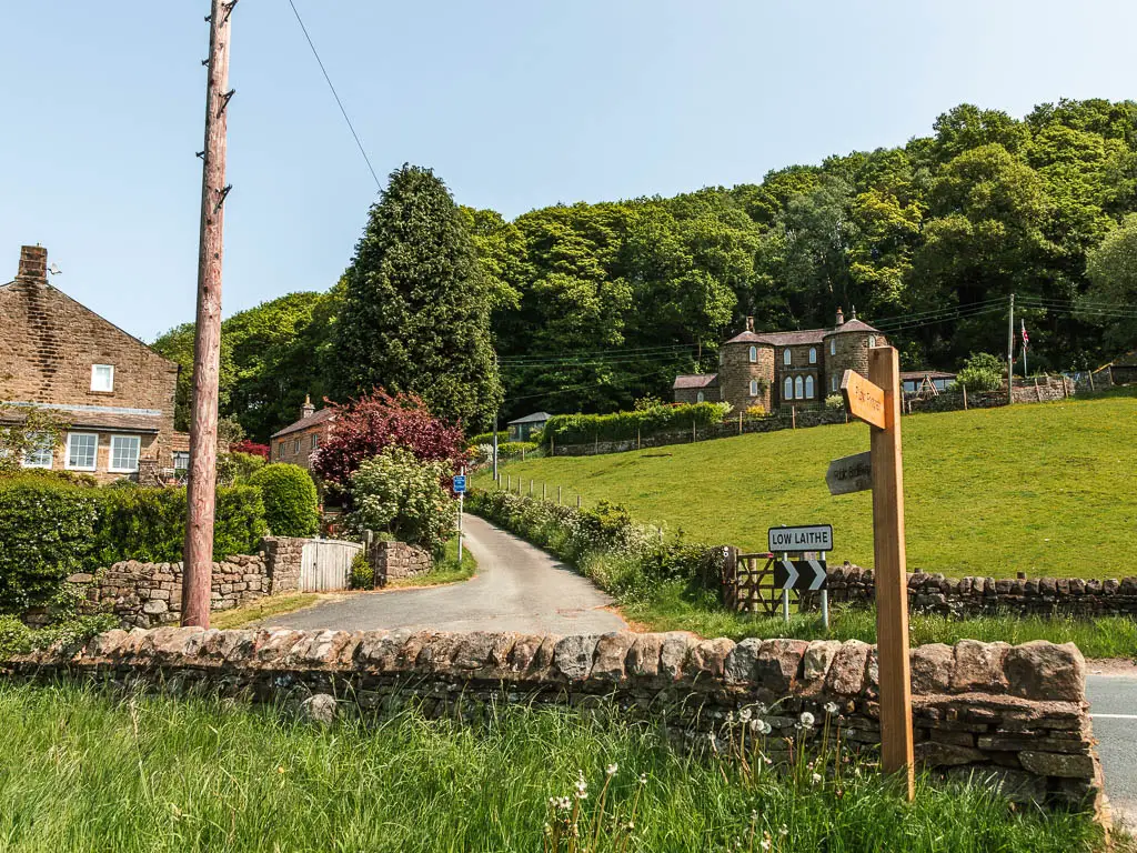 A stone wall, with the road on the other side. There is a side road leading up the hill. There is a wooden trail signpost pointing up the side road. The side road has some voluted bushes on the left side of it, and a grass hills field on the right side. There is a house at the top of the right sided field, with a mass of green leaved trees behind it.