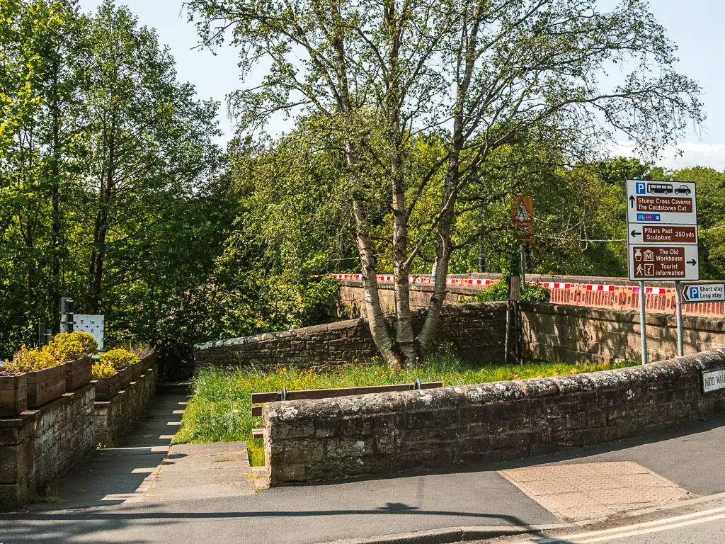 A pavement, lined with a stone wall, and a gap in the wall leading to the river and trees.