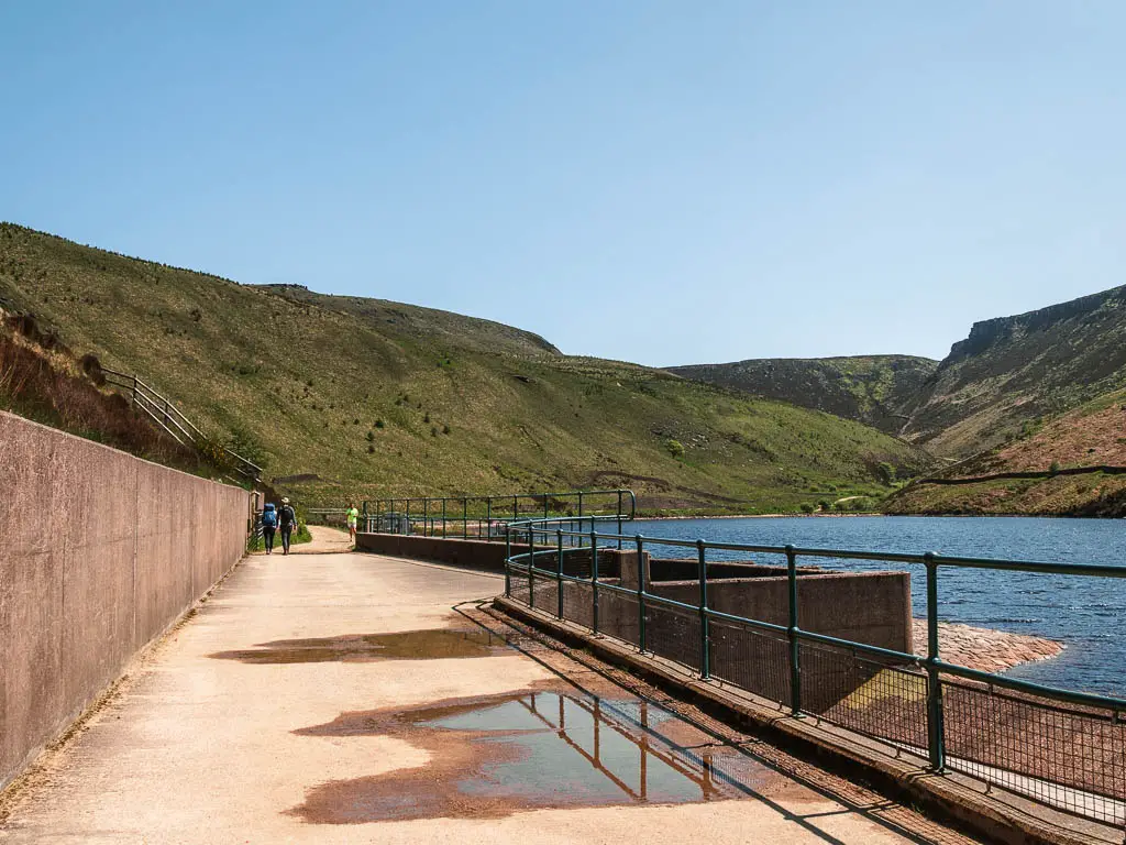 A wide path with a railing on the right, and the reservoir on the other side, with a wall of green hills ahead. 