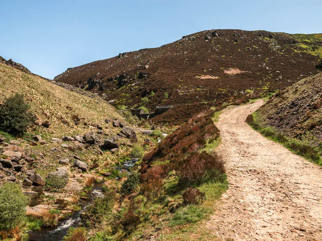 A gravel dirt trail curving uphill on the right, with a small drop to the left, to a small stream of water.