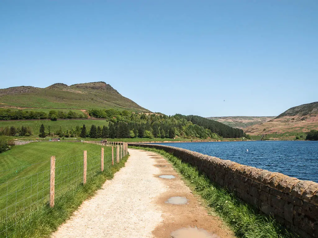 A wide well maintained path with a stone wall and the blue water of the Dovestone reservoir to the right, and a wire fence and green downhill to the left. There is a big green grass covered hill ahead in the distance, with trees below it.