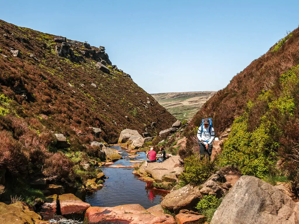 A pool of water surrounded by rocks, sitting in the narrow valley, on the hike up to the Trinnacle. There is a person with a white top walking along the trail on the right, and a couple of people sitting on a rock by the pool of water.