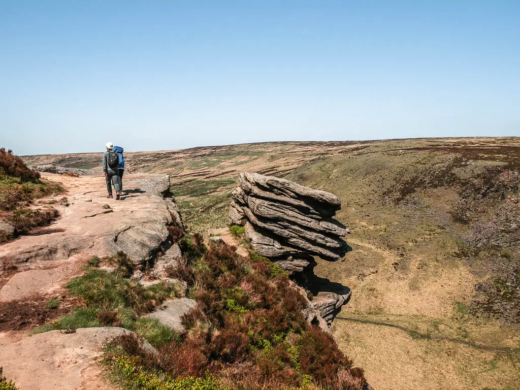 A big rock platform on the edge of the hill with a big rock formation leaning off the side, on the walk to the Trinnacle. There are a couple of people walking along the rock platform ahead.