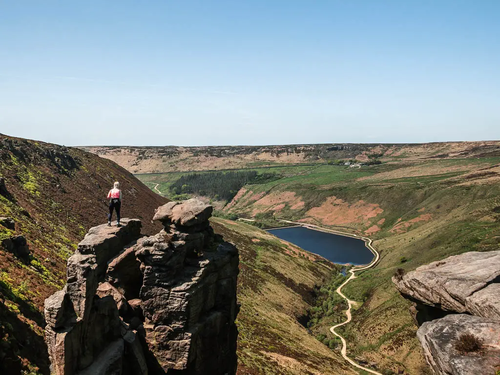 The big Trinnacle rock on the left, with a view down into the valley and trail leading to the blue reservoir below. There is a person standing on top of the Trinnacle.