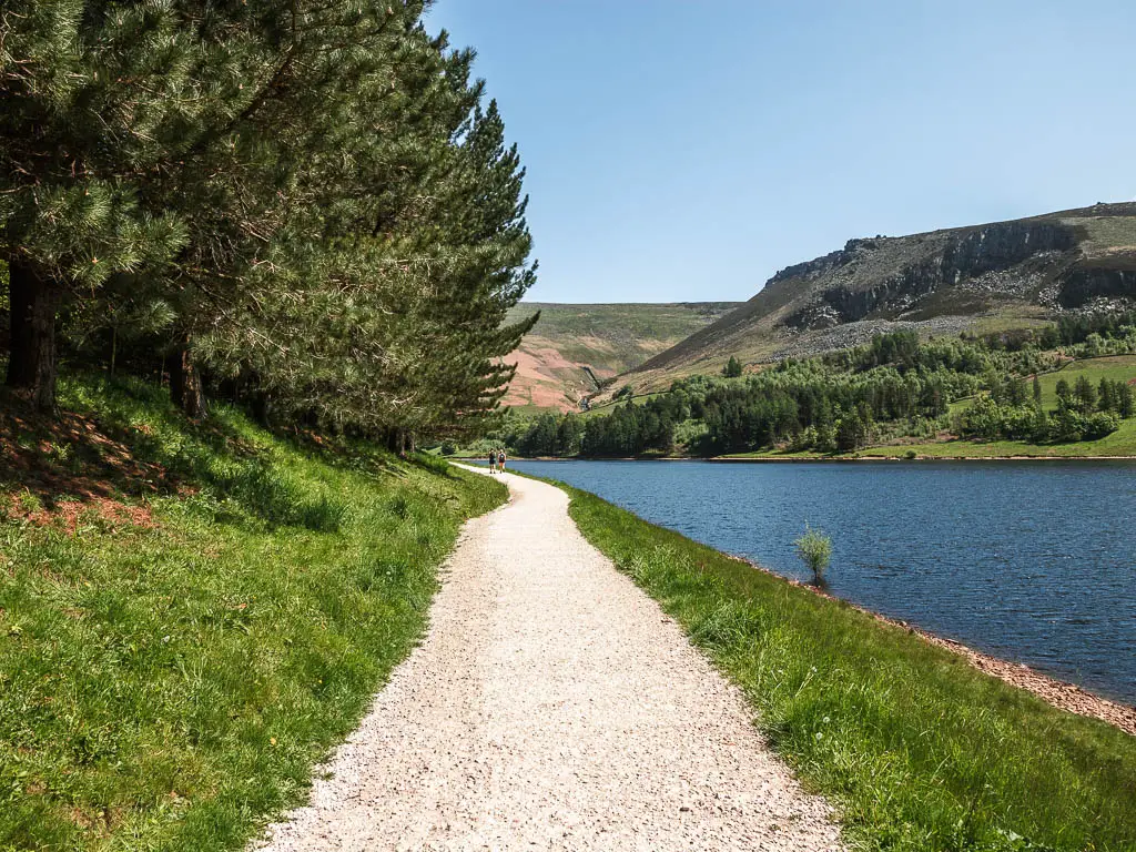 A wide gravel white path, lined with green grass banks, and the blue water of the Dovestone Reservoir to the right, and trees up the bank to the left.