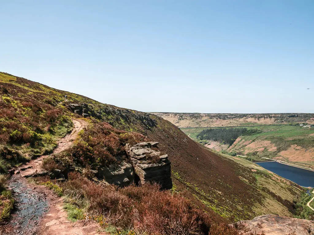 A dirt trail on the left, along the side of the steep hill, on the walk away from the Trinnacle. There is a view down into the valley to the right, with the reservoir at the bottom.