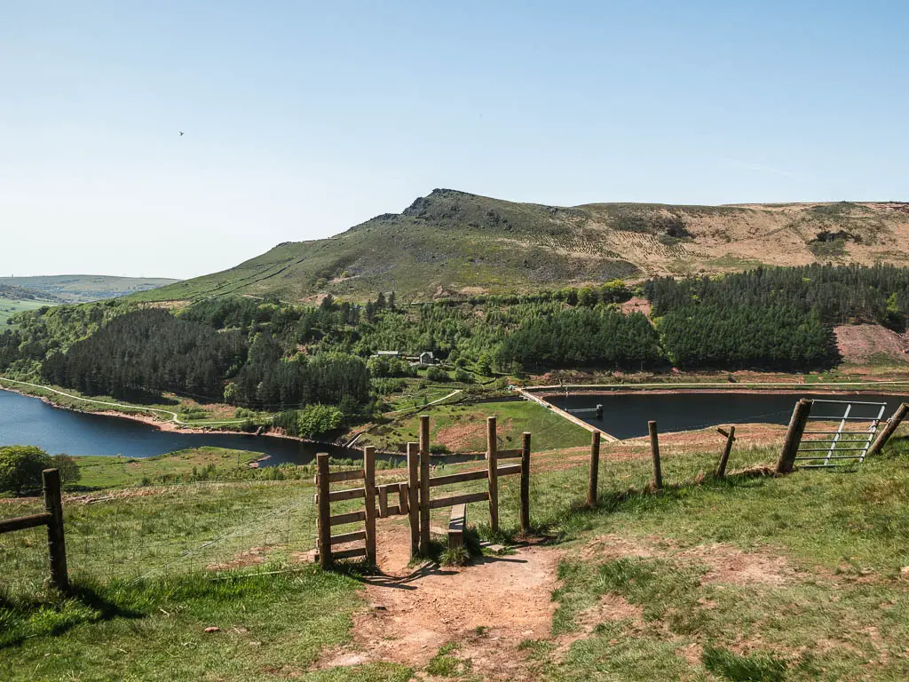 A fence and wooden stile halfway down the grass hillside, with the two reservoirs below, and a mass of trees and big hill on the other side of the water.