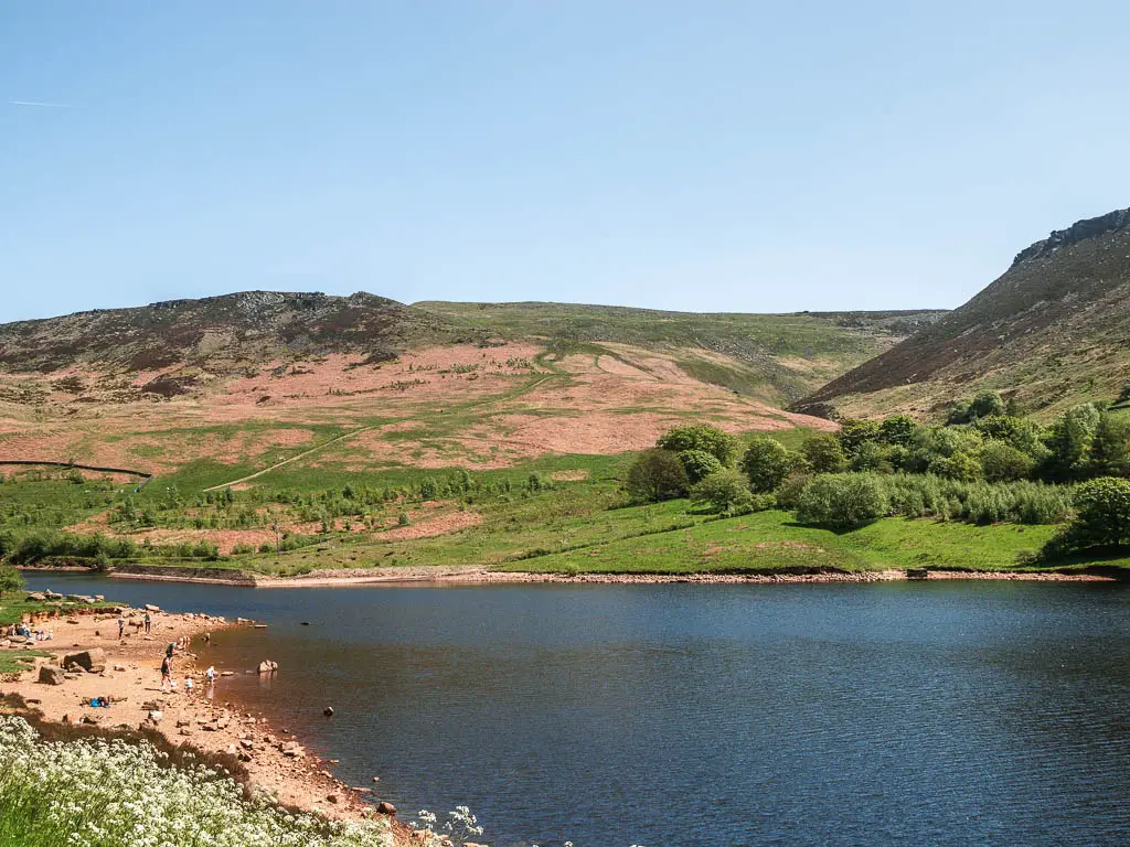 Looking across the blue water of the Dovestone Reservoir, with hills rising up on the other side, on the walk towards the Trinnacle. There is a small sand bank on the left, with a few people walking on it. 