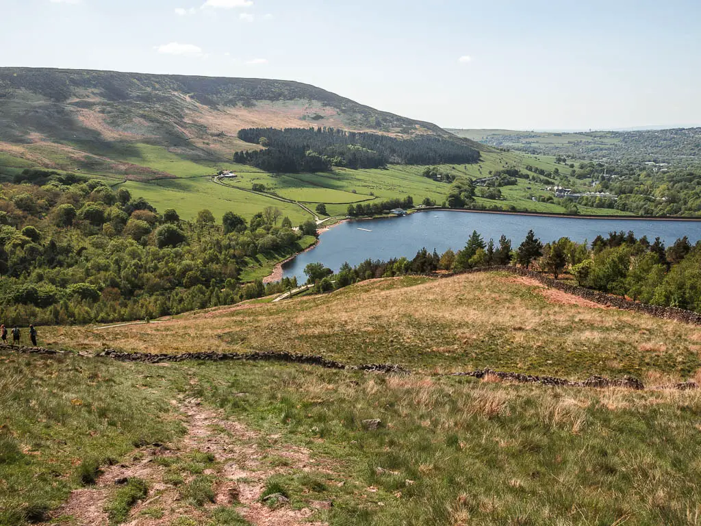 A dirt trail leading down the grass hill, near the end of the Trinnacle circular walk. The reservoir is at the bottom of the hill, with a mass of woodland trees to the left, and grass fields and hill rising up on the other side.