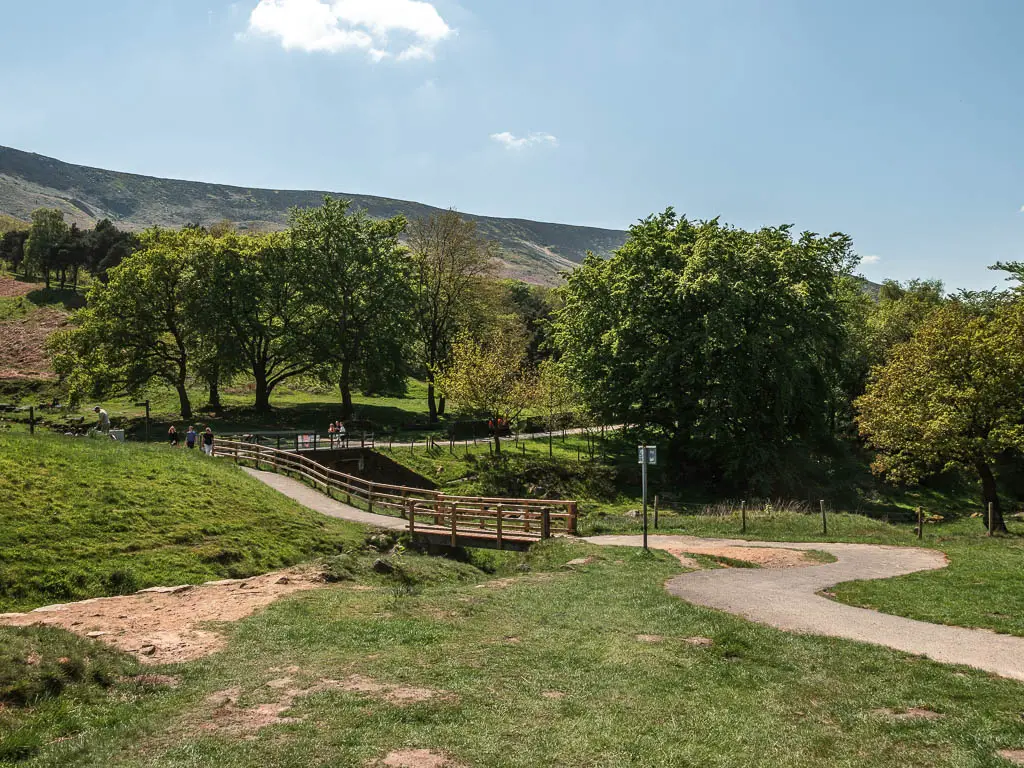 A winding path running along the right side of the grass area, with railings lining the path head. There is a mass of trees on the other side.