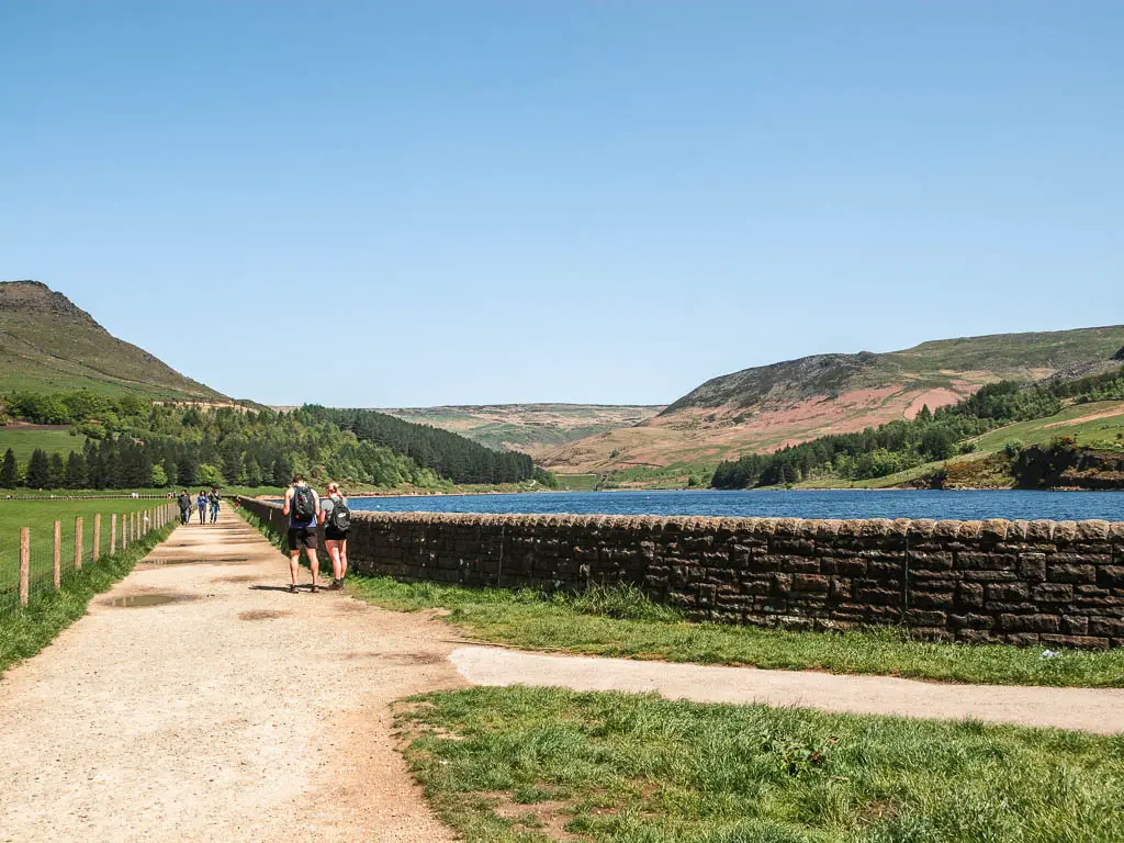 The wide well maintained path running around the corner of the Dovestone Reservoir, which is lined with a stone wall. There are people walking along the path.