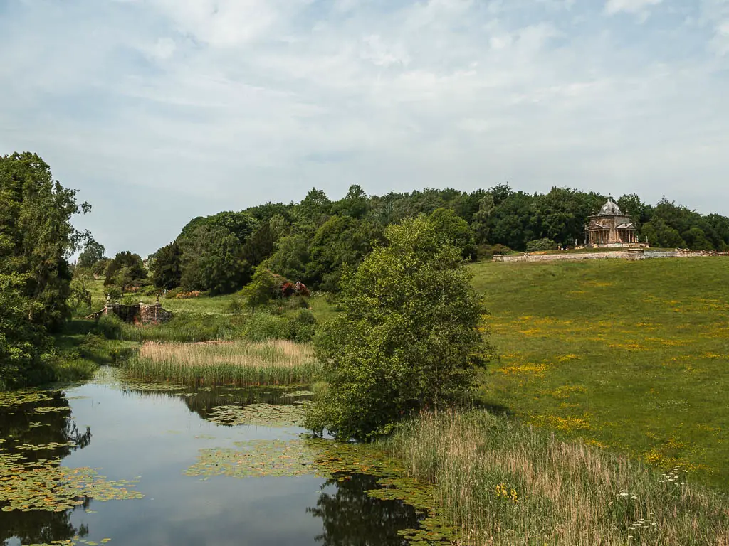 A pond on the left, and a grass hill field on the right, with the Temple of the Four winds on top of the hill, on the Castle Howard circular walk.