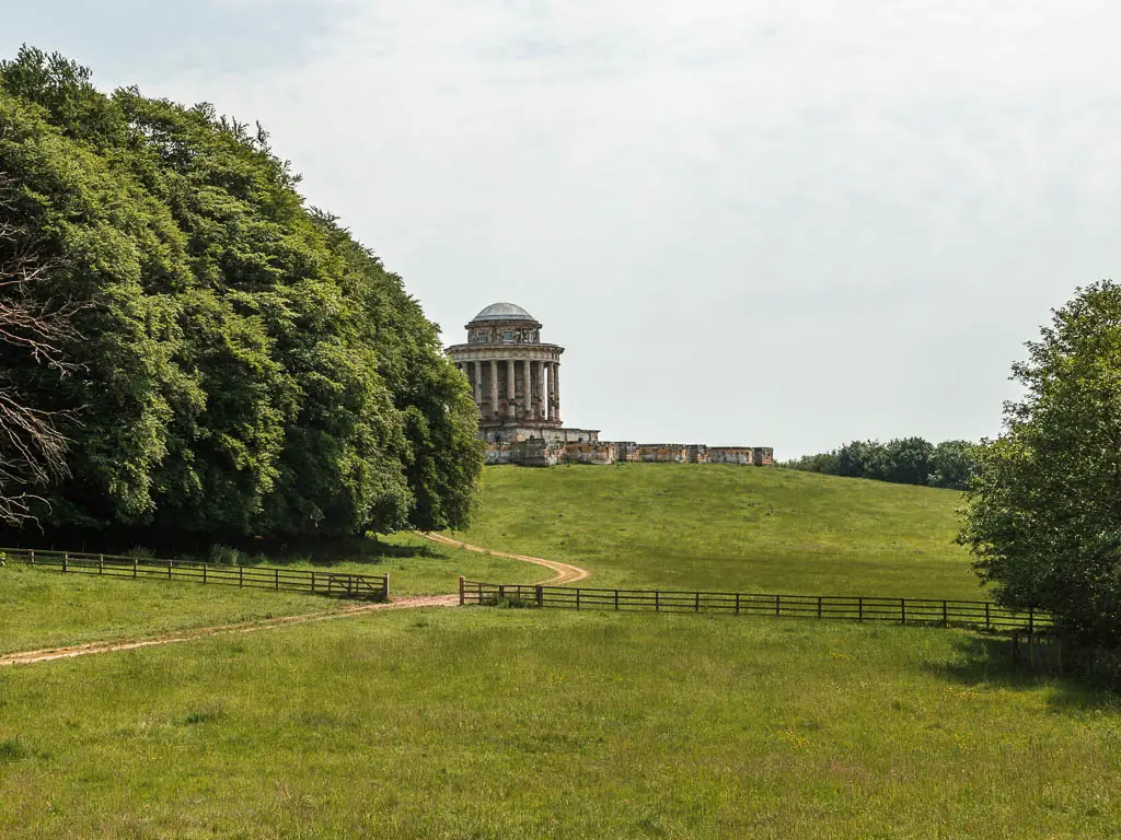 Looking across the grass field, towards the Castle Howard Mausoleum on the hill ahead, partway through the walk. There is a mass of green leafy trees to the left.