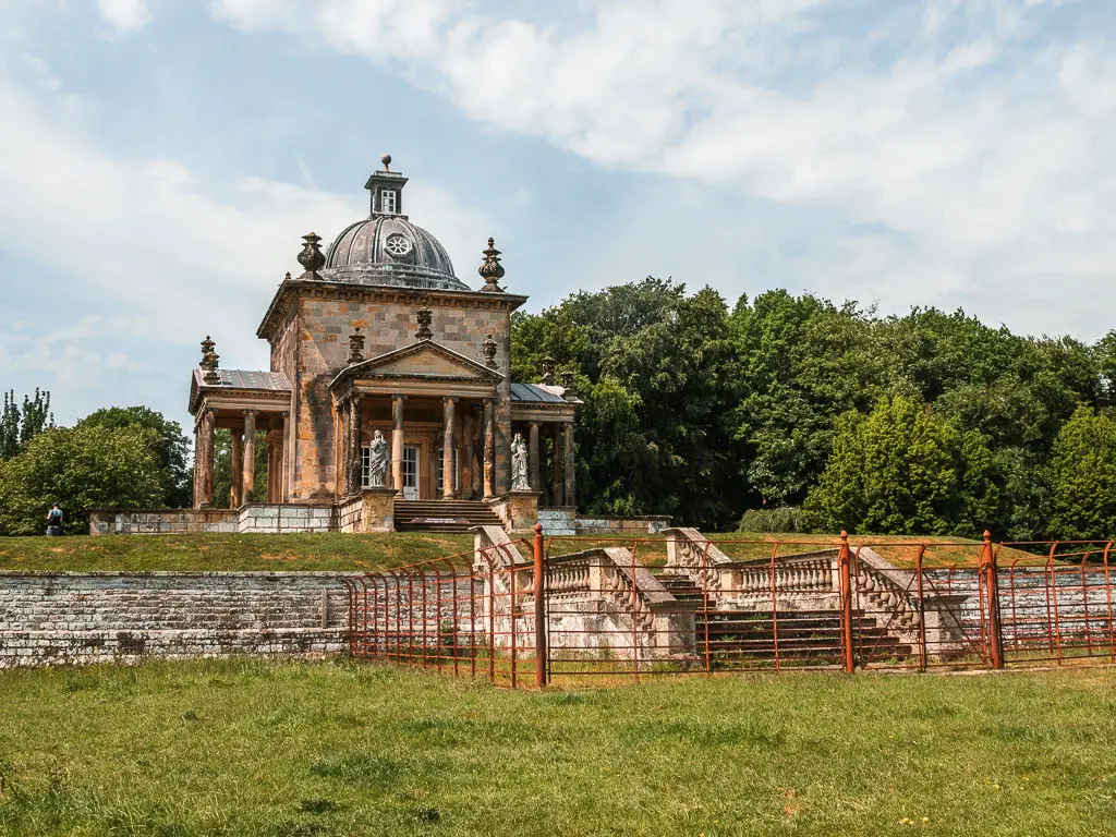 The Temple of the Four winds, sitting on the hill too, surround by a stone wall and red iron fence, on the Castle Howard circular walk. There are green leaved trees behind the temple.