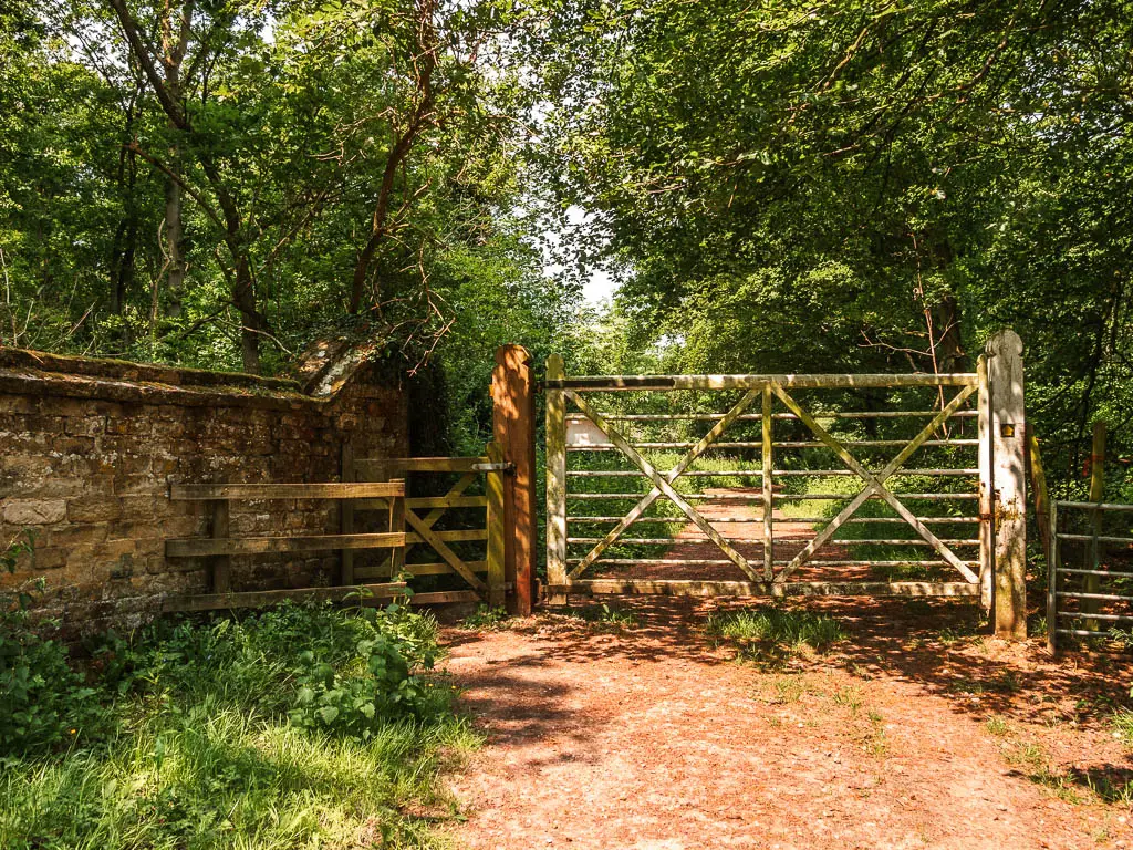 The path leading though a small wooden gate and big metal gate, with a stone wall to the left, and woodland on the other side.