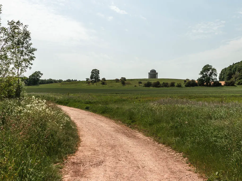 A dirt road curving to the left, with grass fields to the right, and the Castle Howard Mausoleum on the hill in the distance, partway through the walk.
