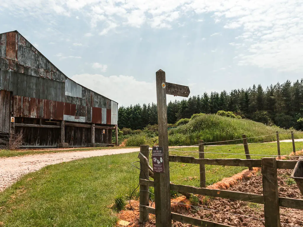 A wooden trail signpost on the corner of a wooden fence. It is pointing right, along the gravel path. There is a big farm building on the other side of the path on the left.