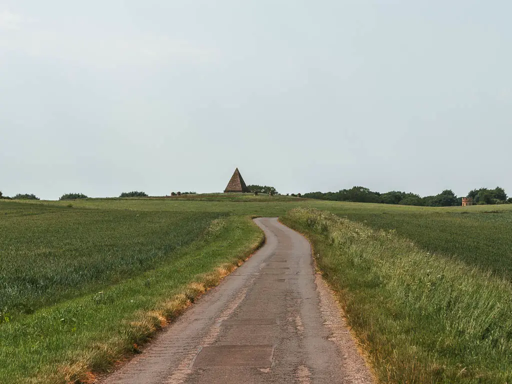 A road leading straight ahead, surround by green fields, with a pyramid ahead in the distance.