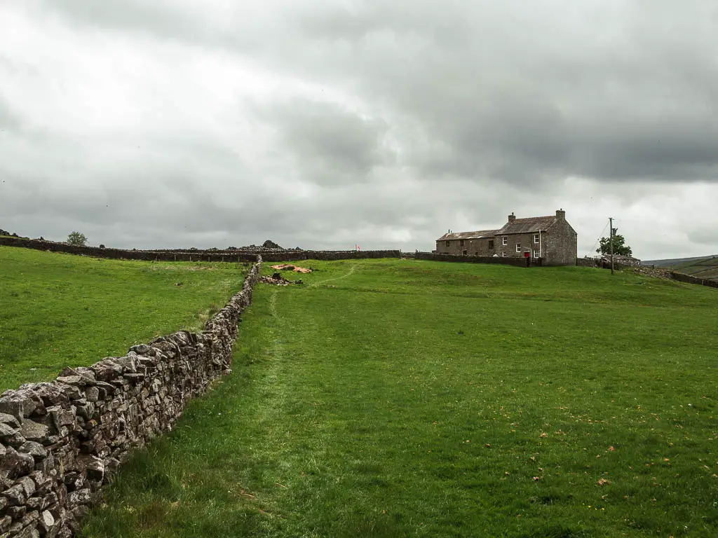 A stone wall running straight head across a large grass field, partway through the Fremington Edge circular walk. There is a stone walled farm house on the other side of the field.