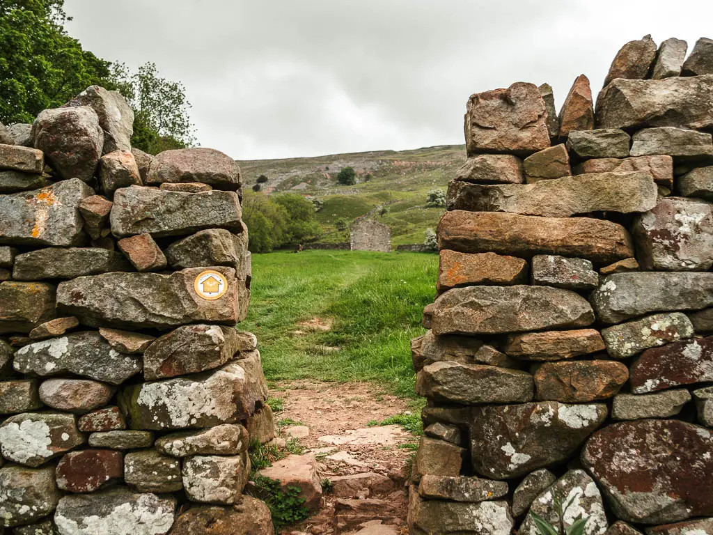 Looking through a gap in the stone wall. A stone shed is visible through the gap, in the green grass field on the other side. There is a yellow arrow on the stone wall, pointing through it.