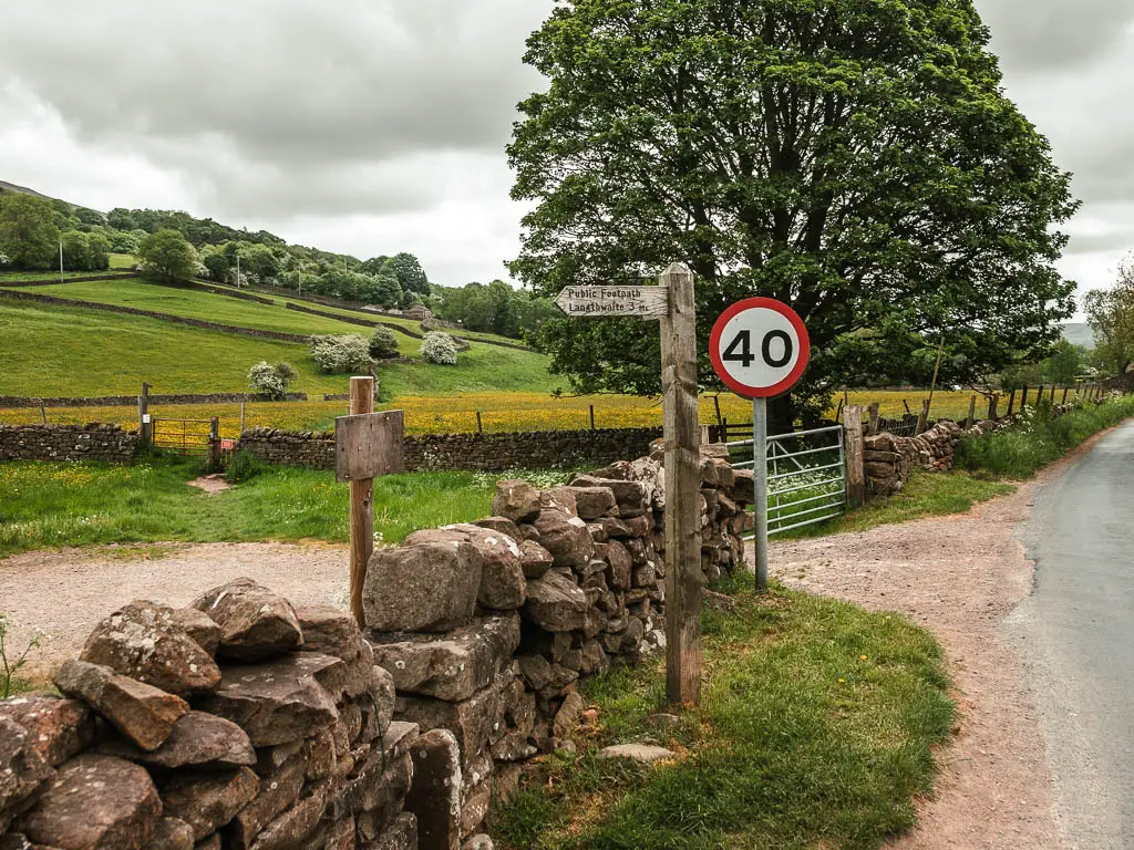 A stone wall, with a wooden trail signpost pointing left over it. There are hills with trees dotted about ahead. The road is to the right.