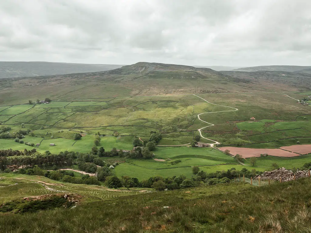 Looking down into the valley on the Fremington Edge walk. The valley is filled with green fields, with a hill and peak rising up on the other side, with a road curving up it.
